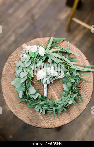 Mani rendendo ghirlanda di nozze da eucalypthus e fiori e boutonniere per lo sposo da fiori di colore bianco e verde sul tavolo di legno sfondo rustico, Foto Stock