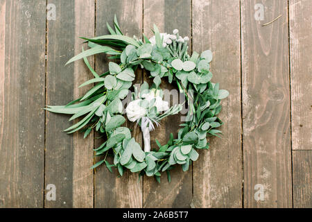 Mani rendendo ghirlanda di nozze da eucalypthus e fiori e boutonniere per lo sposo da fiori di colore bianco e verde su sfondo di legno, decorazioni rustiche Foto Stock