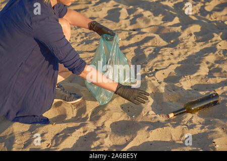 Giovane donna in nero guanti è camminare con il sacchetto di immondizia lungo una spiaggia sporca del fiume e per ripulire il cestino, utilizzate bottiglie in vetro. Costa dell'inquinamento. V Foto Stock