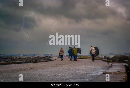 Un gruppo di persone che camminano e cercando di mantenere gli ombrelli in una giornata di vento e fulmini in background Foto Stock