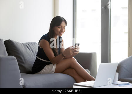 Fiducioso Asian imprenditrice utilizzando phone, seduto sul lettino in ufficio Foto Stock