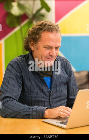 Un uomo si siede a un tavolo in una sala colorata al lavoro su un notebook Foto Stock