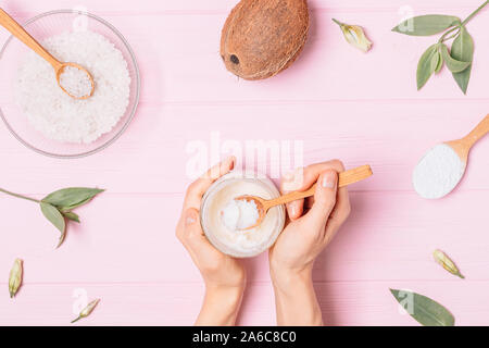 Vista superiore mani femminili tenendo un vasetto di olio di noce di cocco con il cucchiaio di legno accanto a ingredienti cosmetici sale marino e argilla bianca su sfondo rosa, piatto laici. Foto Stock