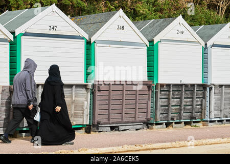 Una donna in nero pieno hijab nikab passeggiate lungo la passeggiata con suo marito passato cabine sulla spiaggia, accanto alla spiaggia di sabbia a Bournemouth, Inghilterra. Foto Stock