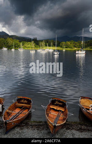 Velieri ormeggiati sul lago di Windermere con imbarcazioni a remi sul litorale di Waterhead Ambleside nel sole di mattina con nuvole scure Parco Nazionale del Distretto dei Laghi Foto Stock