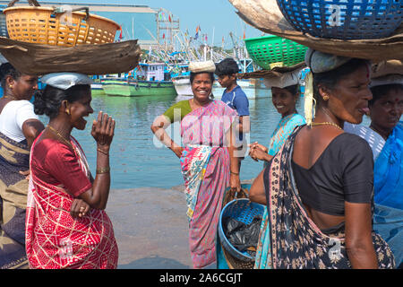 I venditori di pesci femmine che acquistano pesce al vecchio porto di Mangalore, Karnataka, India, si levano in piedi intorno scherzando, con le barche di pesca nello sfondo Foto Stock