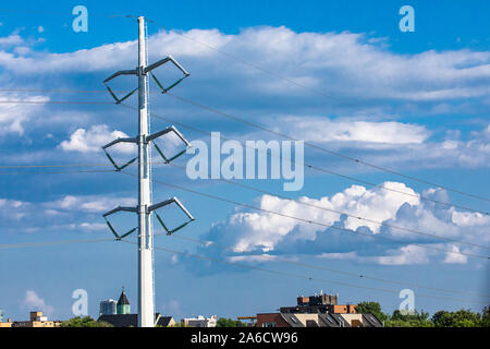 Una vista ingrandita di un bianco pilone elettrico il supporto di linee elettriche aeree in una zona urbana, con tetti contro un cielo blu con nuvole e copia di spazio. Foto Stock