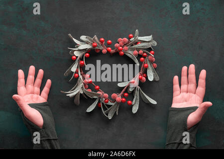 Mani femminili che mostra a forma di cuore vischio decorativa ghirlanda di Natale smerigliato con foglie e bacche di colore rosso. Appartamento laici scuro su sfondo a trama Foto Stock