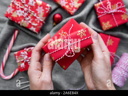 Sfondo di natale con confezioni regalo, bugne di corda e decorazioni su rosso. Preparazione per le vacanze. Vista dall'alto. Donna di mani confezione regalo. Foto Stock