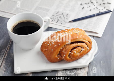 Torta svedese alla cannella e una tazza di caffè nero contro un giornale con crossword Foto Stock