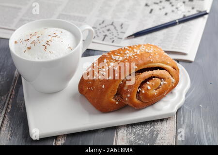 Torta svedese alla cannella e una tazza di cappuccino contro un giornale con crossword Foto Stock