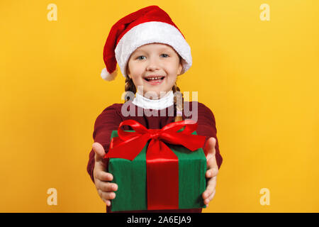 Bambino felice con il regalo di Natale. Bambina in santa claus hat offre un regalo su uno sfondo giallo. Foto Stock