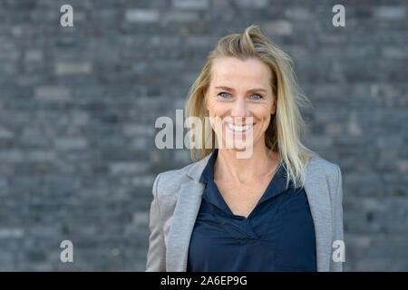 Ritratto di sorridere donna bionda con i capelli dritti, indossando il blu scuro camicetta e rimosse blazer grigio in piedi all'aperto contro sfocata backgro grigio Foto Stock