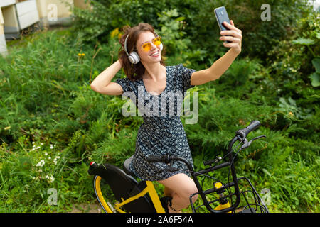 Giovane donna caucasica con Perfect Smile, labbra carnose, bicchieri, auricolari, passeggiate nella natura, un giro in bici e registra un video, fare selfie Foto Stock