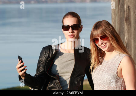 Due ragazze con occhiali da sole dal lago tenendo selfies e divertirsi Foto Stock