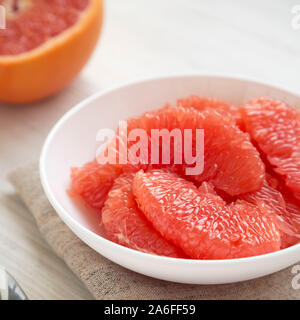 Pompelmo rosso fette in una ciotola bianco su bianco di una superficie di legno, vista laterale. Close-up. Foto Stock