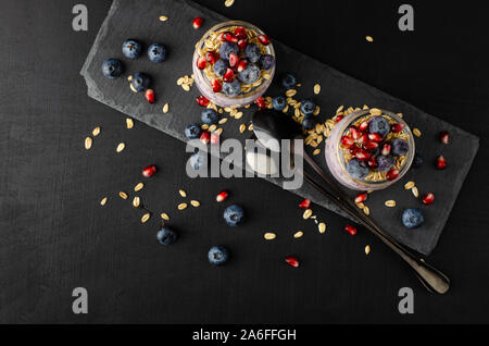 Fitness sano concetto di cibo. I vasetti di yogurt con mirtilli, i semi di melograno e avena su sfondo nero. top view, laici piatta con spazio di copia Foto Stock