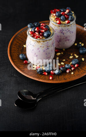 Colazione sana nozione. Vasetti di yogurt fatto in casa con mirtilli, i semi di melograno e avena sull'argilla rossa piastra. Verticale. Foto Stock