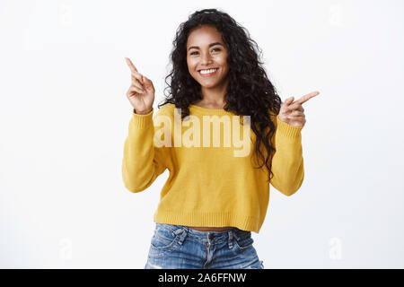 Allegro amichevole-cercando attraente sorridente AFRICAN-AMERICAN girl in maglione giallo rivolto lateralmente, indicando due lati, pubblicità links Foto Stock