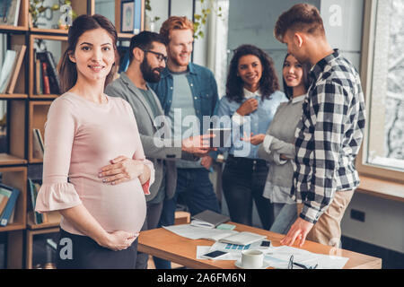 Startupers lavorando insieme a Office donna incinta close-up telecamera cercando felice mentre i colleghi a discutere di progetto Foto Stock