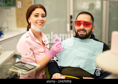 Giovani femmine dentista medico e paziente sorridente in telecamera presso la clinica dentale. Occhiali di sicurezza per il paziente. Il medico in indumenti protettivi personali equi Foto Stock