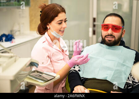 Giovani femmine dentista medico e paziente sorridente in telecamera presso la clinica dentale. Occhiali di sicurezza per il paziente. Il medico in indumenti protettivi personali equi Foto Stock