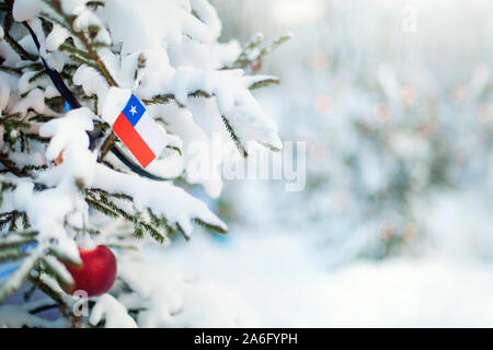 Natale Cile. Albero di natale con neve, decorazioni e una bandiera del Cile. Boschi innevati background in inverno. Natale biglietto di auguri. Foto Stock