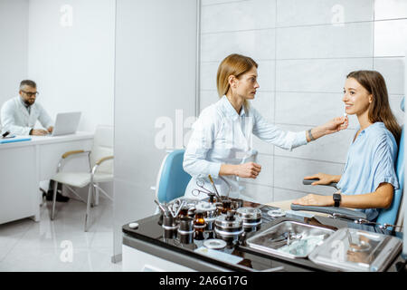 Otolaryngologist femmina esaminando le orecchie con ENT diapason per un paziente giovane nell'ufficio medico con medico senior sullo sfondo Foto Stock