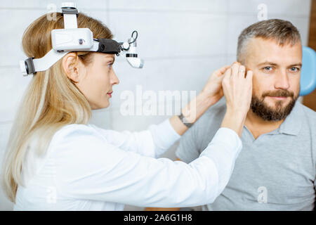 Otolaryngologist femmina con paziente adulto durante un esame medico di un orecchio alla stanza di procedura dell'ufficio ENT Foto Stock