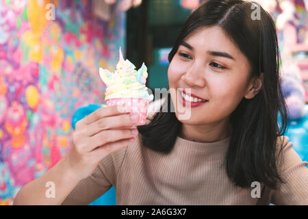 Felice giovane americano ragazza asiatica, mangiare multicolor unicorn cupcake in città alla moda cafe - moda, stile di vita alimentare i concetti di blogger Foto Stock