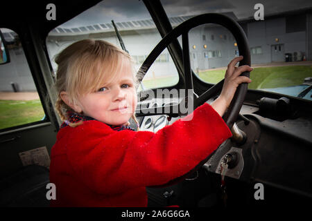 Un incantevole piccola ragazza bionda in un cappotto rosso tiene il volante di un camion vintage Foto Stock