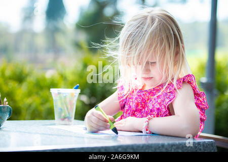 Un simpatico bambina indossa un abito grazioso gode facendo alcune colorazioni in attesa per la sua cena per arrivare, mangiare all'aperto sotto il sole Foto Stock