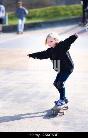 Un grazioso piccolo bionda ragazza dai capelli sul suo skateboard di praticare il pattinaggio presso il locale skatepark, 7 anni Foto Stock