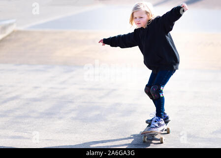 Un grazioso piccolo bionda ragazza dai capelli sul suo skateboard di praticare il pattinaggio presso il locale skatepark, 7 anni Foto Stock