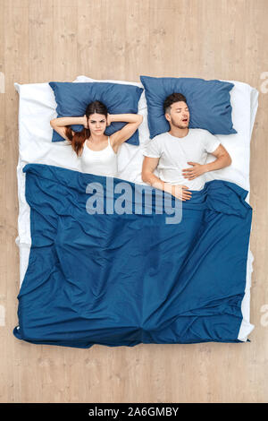 Andare a dormire. Giovane uomo dorme sbadigliare mentre la donna che copre le orecchie arrabbiato sotto la coperta sul letto vista superiore Foto Stock
