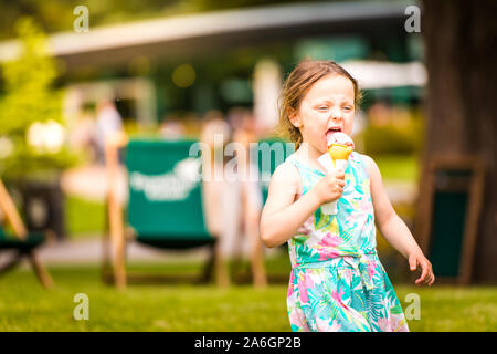 Un simpatico bambina in una luminosa abiti estivi di mangiare un gelato e giocare sulle altalene a Trentham Gardens in una calda giornata d'estate Foto Stock