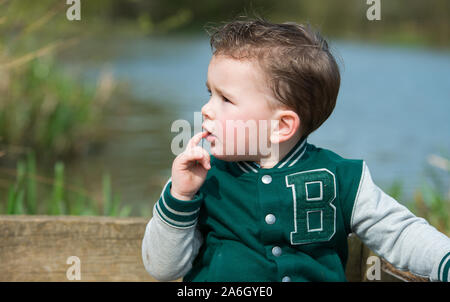 Un grazioso piccolo ragazzo seduto in riva al lago a Colchester Country Park, Essex Foto Stock