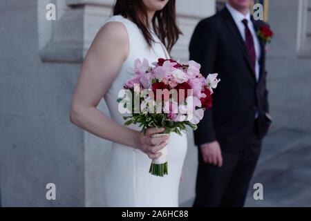 Closeup incinta sposa in abito bianco bouquet di contenimento del rosso e del rosa rose, lo sposo in un vestito nero accanto a lei, impostazione all'aperto Foto Stock
