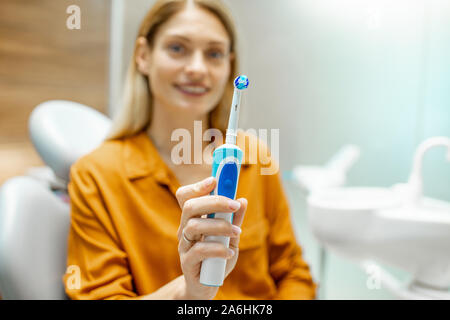 Ritratto di una bellissima e allegra donna come un paziente con spazzolino da denti elettrico seduto sulla poltrona odontoiatrica in studio dentistico. Immagine focalizzata sulla spazzola Foto Stock