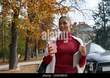Bellissima giovane donna camminare tenendo il suo computer portatile e un drink in una tazza monouso Foto Stock