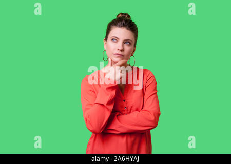 Necessità di pensare! Ritratto di pensieroso elegante giovane donna con bun hairstyle, grandi orecchini e in rosso camicetta mentre tiene il suo mento e seriamente pensando ind. Foto Stock