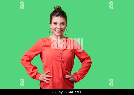 Ritratto di positivo fiducioso giovane donna con bun hairstyle, grandi orecchini e in rosso camicetta in piedi, tenendo le mani sui fianchi, bella sorridente a camer Foto Stock