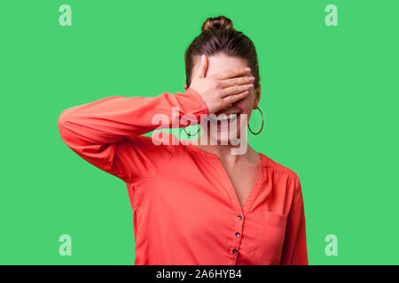 Non si desidera guardare! Ritratto di positivo donna elegante con bun hairstyle, grandi orecchini e in rosso camicetta in piedi che copre gli occhi con la mano e smilin Foto Stock