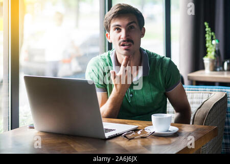 Giovani crazy triste imprenditore in verde t-shirt seduta e di lavoro sul computer portatile con la mano la pistola gun sulla sua testa. business e freelancing problema concetto. Foto Stock