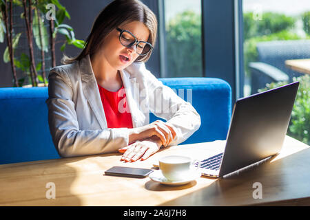 Mano il dolore al polso. Ritratto di triste elegante bruna giovane donna in bicchieri seduta tenendo la sua mano dolorosa dopo un lungo periodo di tempo a lavorare e digitando su laptop Foto Stock