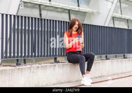 Wow, incredibile news sul telefono cellulare. Ritratto di stupiti affascinante brunette donna in elegante camicia rossa seduta a bocca aperta, messaggio di lettura sulla sm Foto Stock