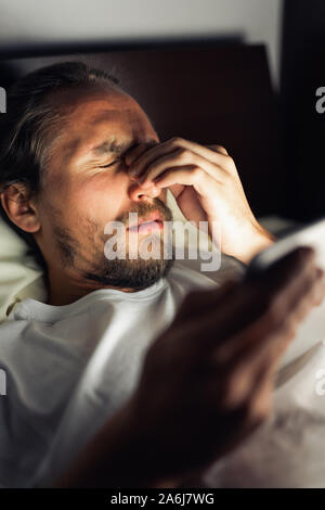 Barbuto maschio caucasico è guardare qualcosa sul suo tablet a letto a notte. Egli è stanco, non riesce a dormire e lui ha chiaramente un mal di testa. Foto Stock