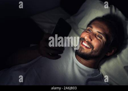 Barbuto felice giovane uomo è sdraiato sul letto e sorridente mentre si guarda qualcosa sul suo telefono. È chiaro che è molto tardi la notte. Foto Stock