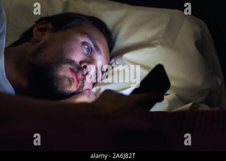 Giovani belli e stanchi con barba non riesce a dormire e guardare qualcosa sul suo telefono cellulare di notte. Foto Stock