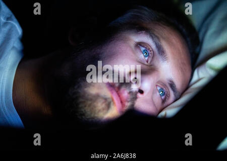 Barbuto maschio caucasico è guardare qualcosa sul suo telefono cellulare a letto a notte. Egli è chiaramente molto stanchi ma non riesce a dormire. Foto Stock